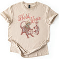 Possum and Frog Comfort Colors T-shirt in Sand, laid flat. A frog in a cowboy hat and bandana rides a snarling possum beside a second possum hanging by its tail, under the words Hold Your in pink script. The shirt lies on a white background with cuffed sleeves.