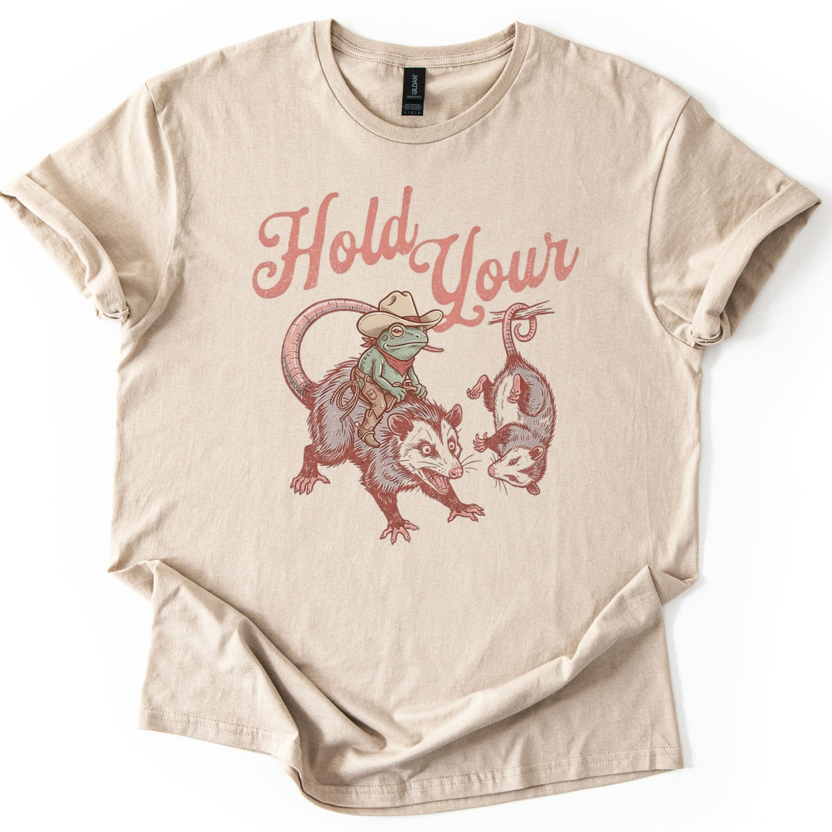 Possum and Frog Comfort Colors T-shirt in Sand, laid flat. A frog in a cowboy hat and bandana rides a snarling possum beside a second possum hanging by its tail, under the words Hold Your in pink script. The shirt lies on a white background with cuffed sleeves.
