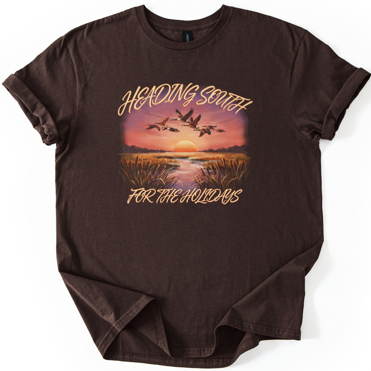 Heading South For The Holidays Comfort Colors T-shirt in Dark Chocolate, laid flat. A flock of geese flies over a marsh with cattails at sunset, between the script words Heading South and For The Holidays. The shirt lies on a white background with its sleeves cuffed.
