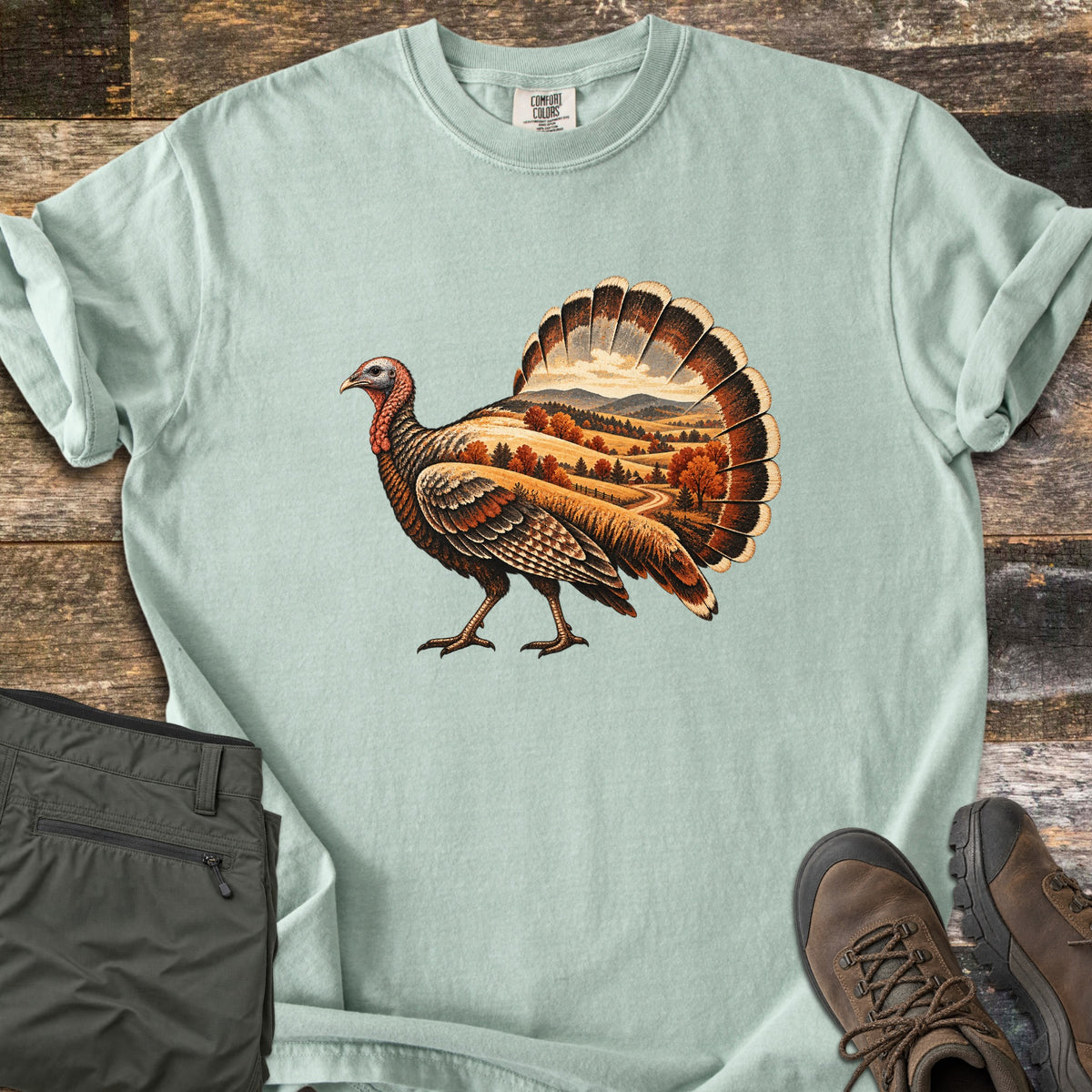 Turkey Harvest Hills Comfort Colors T-shirt in Bay, laid flat. A wild turkey walks left, its body and fanned tail filled with an autumn scene of golden hills, orange trees and a winding road. Sleeves rolled, on weathered wood with olive pants and brown hiking boots.