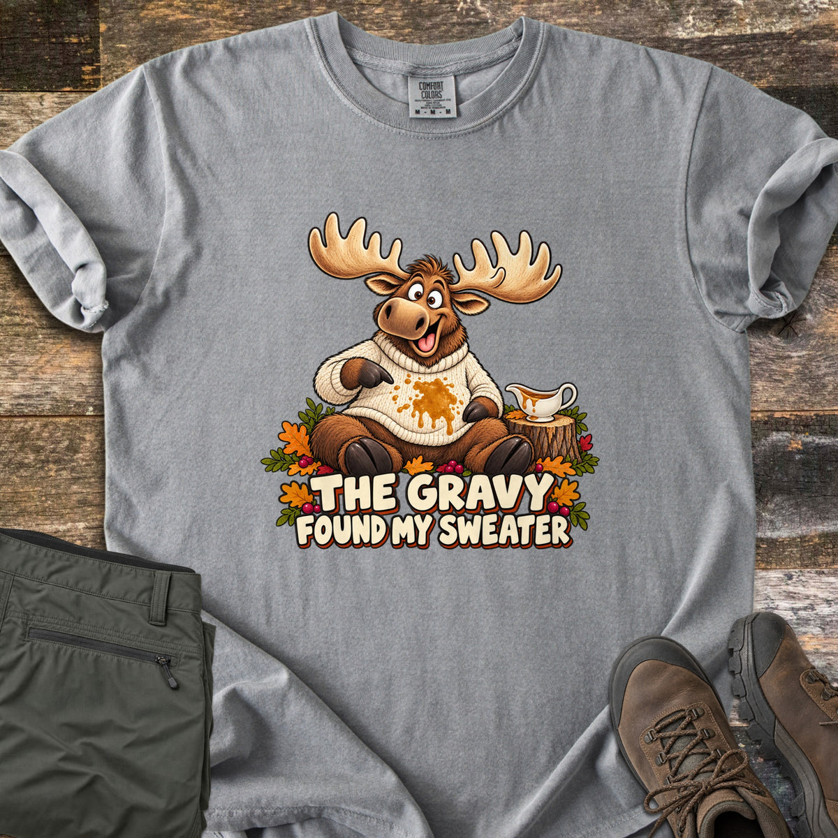 The Gravy Found My Sweater Comfort Colors T-shirt in Grey, laid flat. A cartoon moose in a cream knit sweater points at a gravy stain beside a gravy boat on a stump, above the words The Gravy Found My Sweater. Olive pants and brown boots sit on a wood floor.