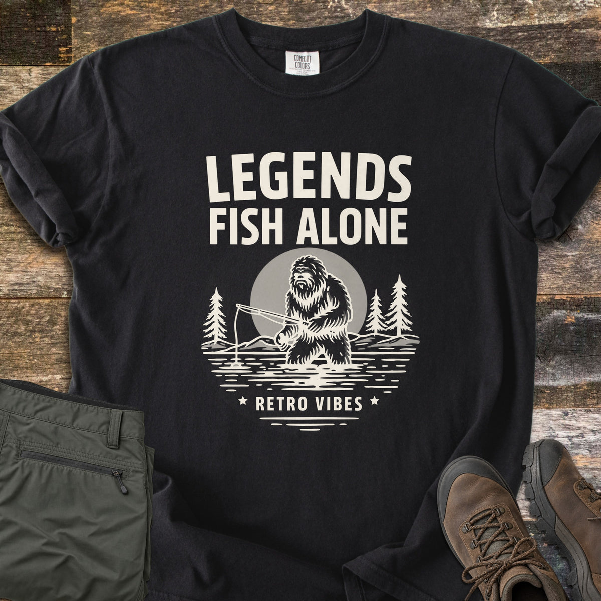 Legends Fish Alone Comfort Colors T-shirt in Black, laid flat. Cream print of the words Legends Fish Alone above a Bigfoot fishing in a lake before a full moon and pines, with Retro Vibes between two stars below. Olive pants and brown hiking boots lie on weathered wood.