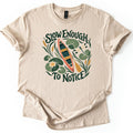 Slow Enough To Notice Comfort Colors T-shirt in Sand, laid flat. A coral canoe with a yellow paddle floats among green lily pads, a water lily, cattails and a dragonfly, framed by the words Slow Enough To Notice in dark green. Sleeves are cuffed, on a white background.