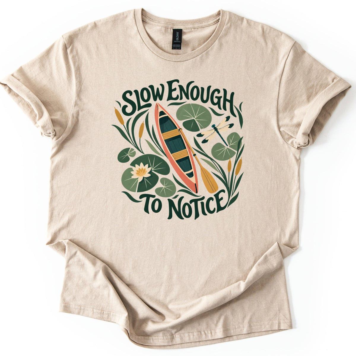 Slow Enough To Notice Comfort Colors T-shirt in Sand, laid flat. A coral canoe with a yellow paddle floats among green lily pads, a water lily, cattails and a dragonfly, framed by the words Slow Enough To Notice in dark green. Sleeves are cuffed, on a white background.
