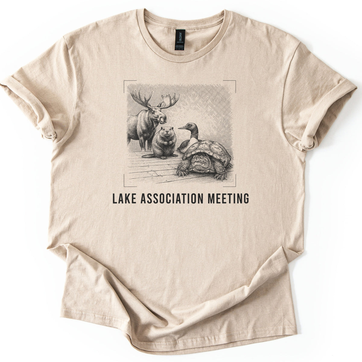 Lake Association Meeting Comfort Colors T-shirt in Sand, laid flat. A black-ink etching of a moose, a beaver, a loon, and a frowning turtle gathered on a wooden dock, framed by corner marks, above the words Lake Association Meeting. Sleeves are cuffed on a white surface.