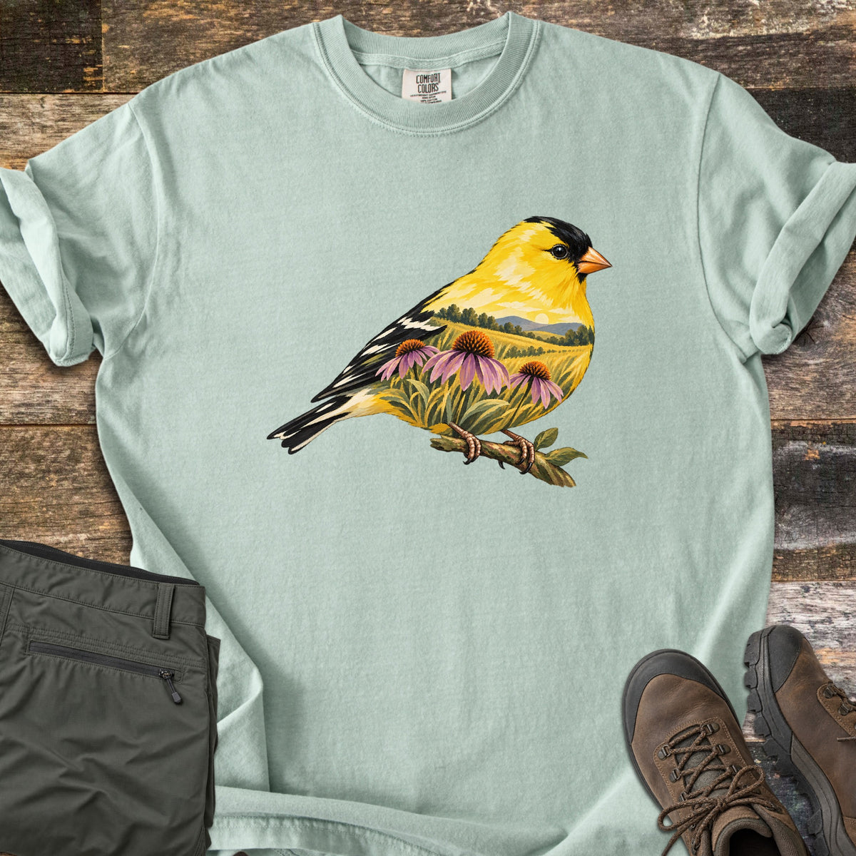 Goldfinch Meadow Comfort Colors T-shirt in Bay, laid flat. A yellow goldfinch perches on a twig, its body filled with a meadow of purple coneflowers, rolling hills and a low sun. No text. Sleeves rolled, beside olive cargo pants and brown hiking boots on weathered wood.
