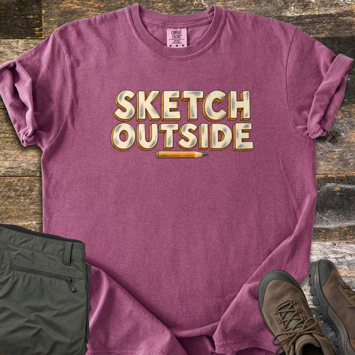 Sketch Outside Comfort Colors T-shirt in Berry, laid flat. The words Sketch Outside in cream block letters with pencil-style shading and orange wood edges sit above a small yellow pencil. Olive hiking pants and brown hiking boots lie beside it on weathered wood planks.