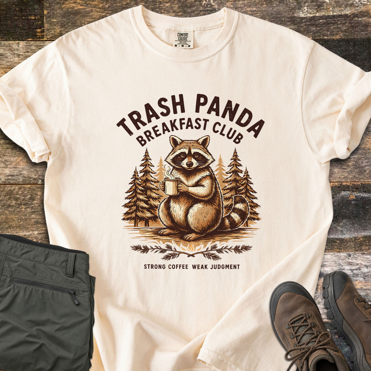 Trash Panda Breakfast Club Comfort Colors T-shirt in Ivory, laid flat. A raccoon holding a steaming mug sits among pine trees under the words Trash Panda Breakfast Club in dark brown, with Strong Coffee Weak Judgment below. Folded green pants and brown hiking boots lie on weathered wood.