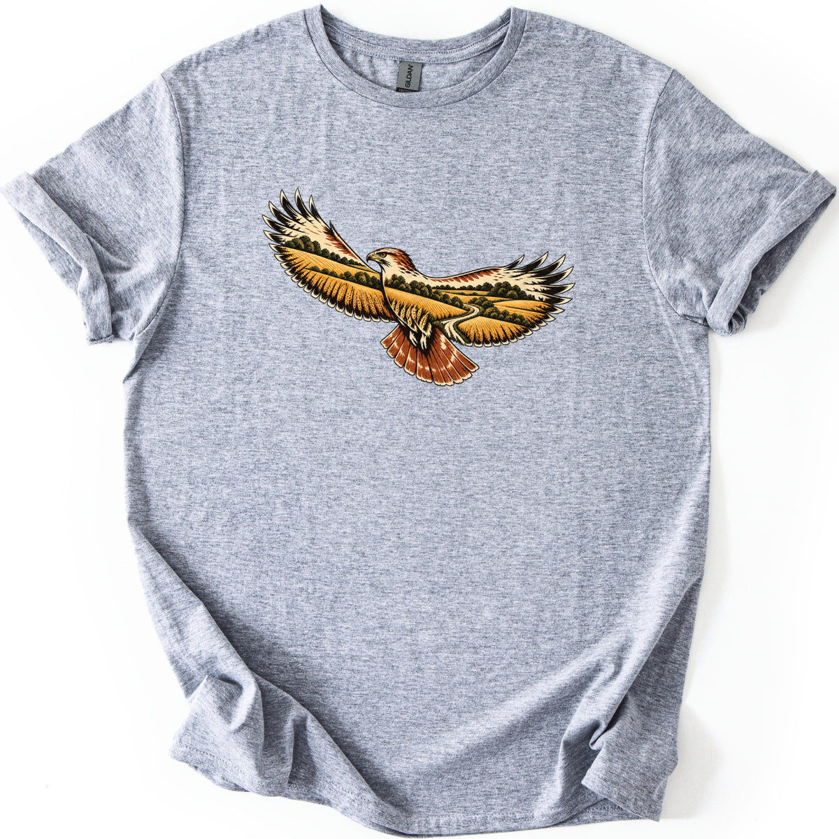 Hawk Over Golden Fields Comfort Colors T-shirt in Sport Grey, laid flat. A soaring hawk with outstretched wings, its wings and body filled with golden wheat fields, green trees and a winding road, and a rust-red tail. No words. The shirt has rolled sleeves on a white surface.