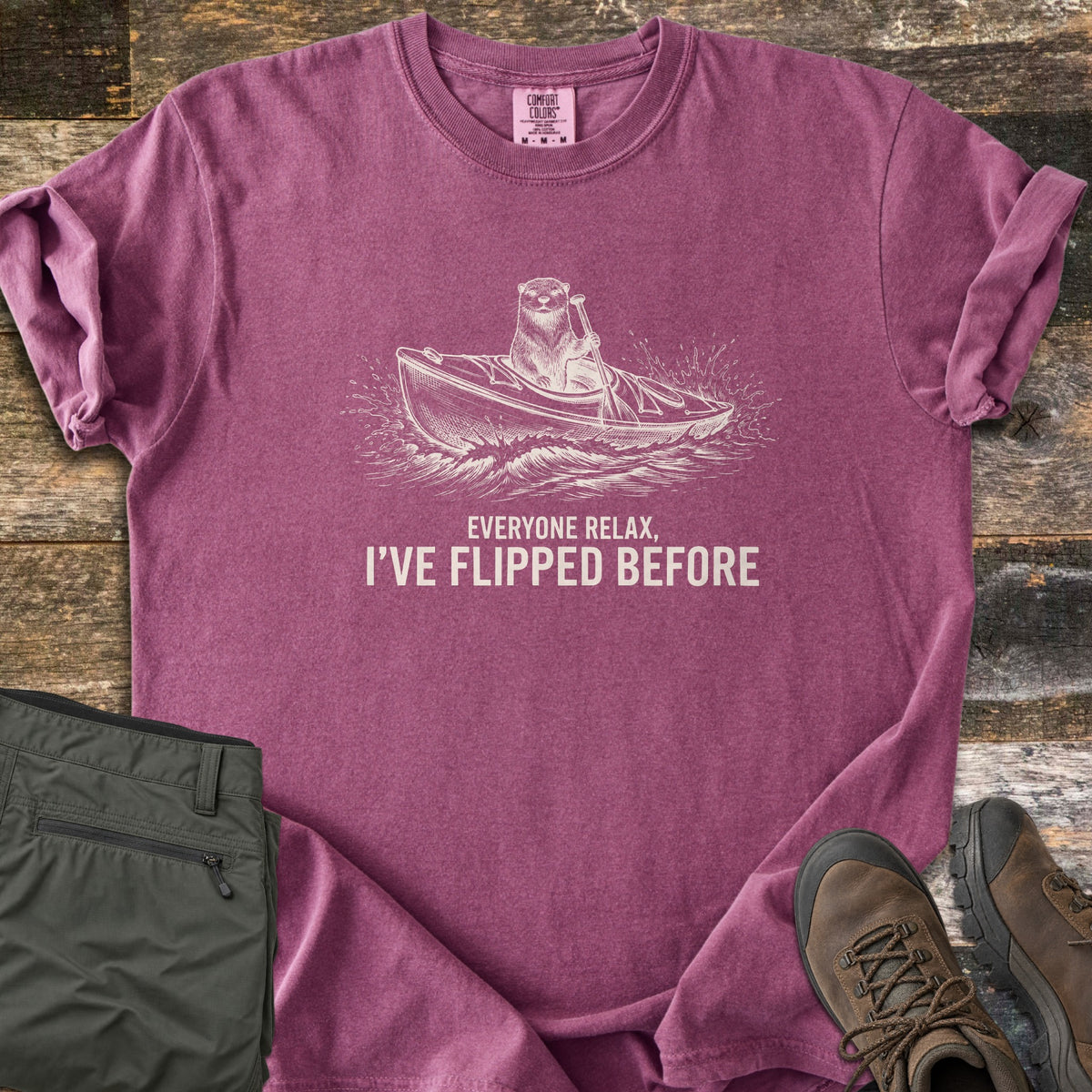 Everyone Relax I've Flipped Before Comfort Colors T-shirt in Berry, laid flat. A cream sketch of an otter paddling a kayak through splashing waves above the words Everyone Relax, I've Flipped Before. Folded olive hiking pants and brown boots lie beside it on weathered wood.