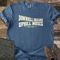 Downhill Dreams Uphill Noises Comfort Colors T-shirt in Denim, laid flat. Cream wavy block letters read Downhill Dreams Uphill Noises above MTB in green between two green lines. Sleeves rolled on weathered wood planks, with folded olive hiking pants and brown hiking boots.