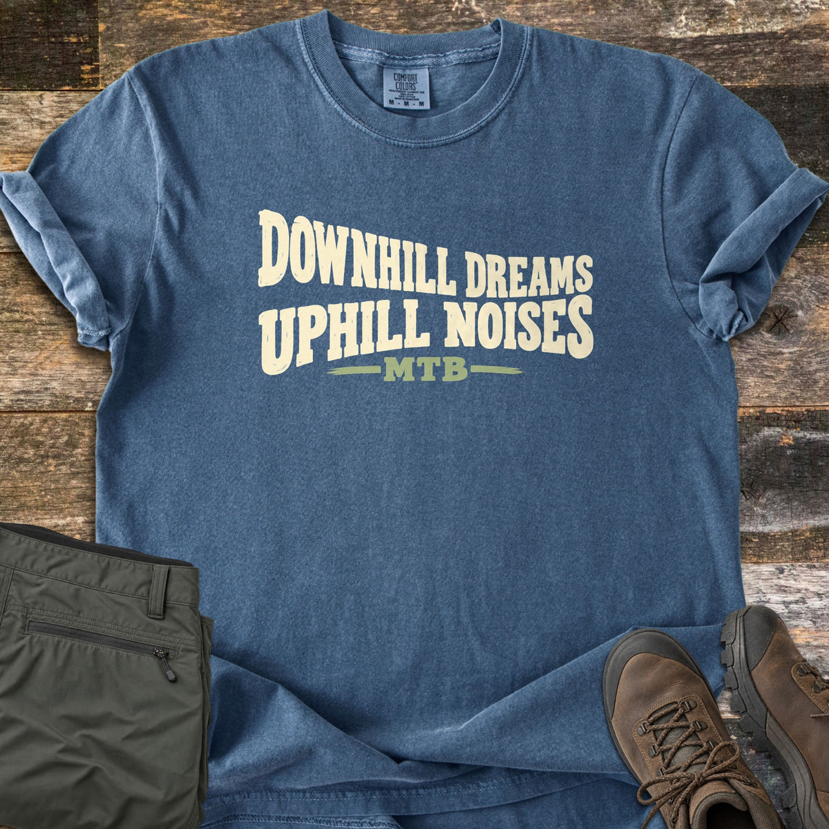 Downhill Dreams Uphill Noises Comfort Colors T-shirt in Denim, laid flat. Cream wavy block letters read Downhill Dreams Uphill Noises above MTB in green between two green lines. Sleeves rolled on weathered wood planks, with folded olive hiking pants and brown hiking boots.