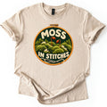Moss in Stitches Comfort Colors T-shirt in Sand, laid flat. An embroidery hoop holds a stitched scene of green moss mounds with two orange spore stalks, the words MOSS IN STITCHES in cream yarn, and a needle with thread. Sleeves rolled, on a white background.