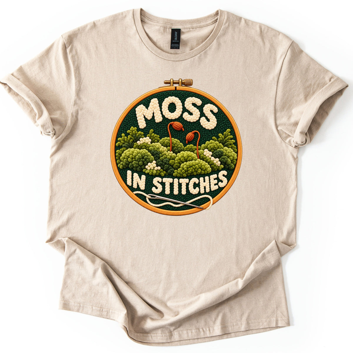 Moss in Stitches Comfort Colors T-shirt in Sand, laid flat. An embroidery hoop holds a stitched scene of green moss mounds with two orange spore stalks, the words MOSS IN STITCHES in cream yarn, and a needle with thread. Sleeves rolled, on a white background.