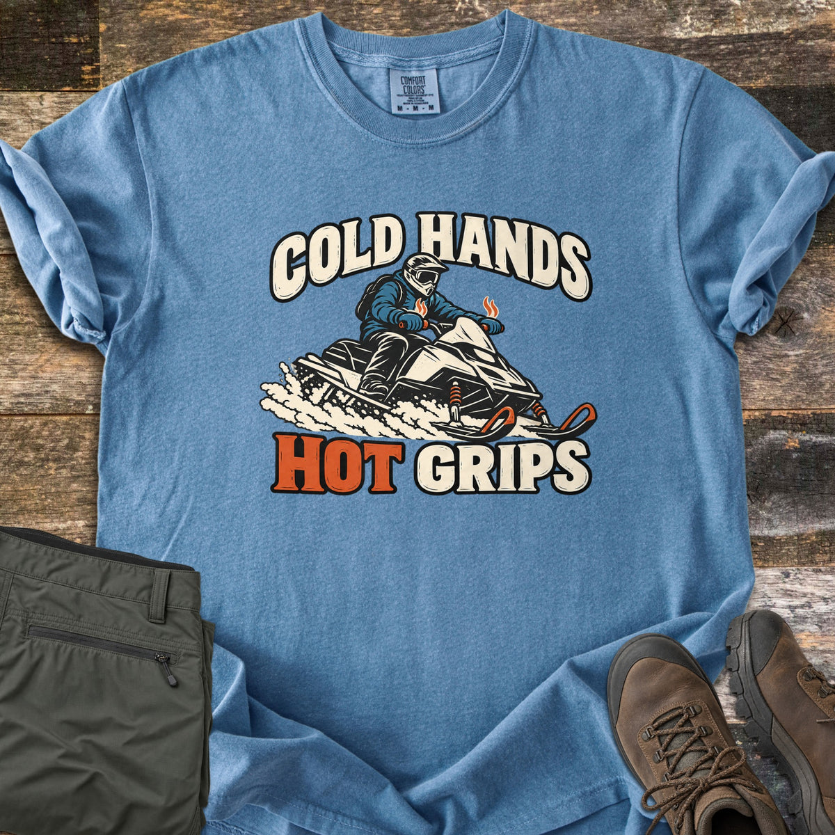 Cold Hands Hot Grips Comfort Colors T-shirt in Blue Jean, laid flat. A snowmobile rider in a helmet and goggles kicks up snow, with steam rising from the handlebar grips, between the words Cold Hands and Hot Grips. Shown on rustic wood with olive cargo pants and brown hiking boots.
