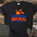 First Break Big Deal Comfort Colors T-shirt in Black, laid flat. An orange clay target shattering into pieces above the words First Break Big Deal Clay Days. Sleeves rolled, on weathered wood with folded olive cargo pants and brown hiking boots.