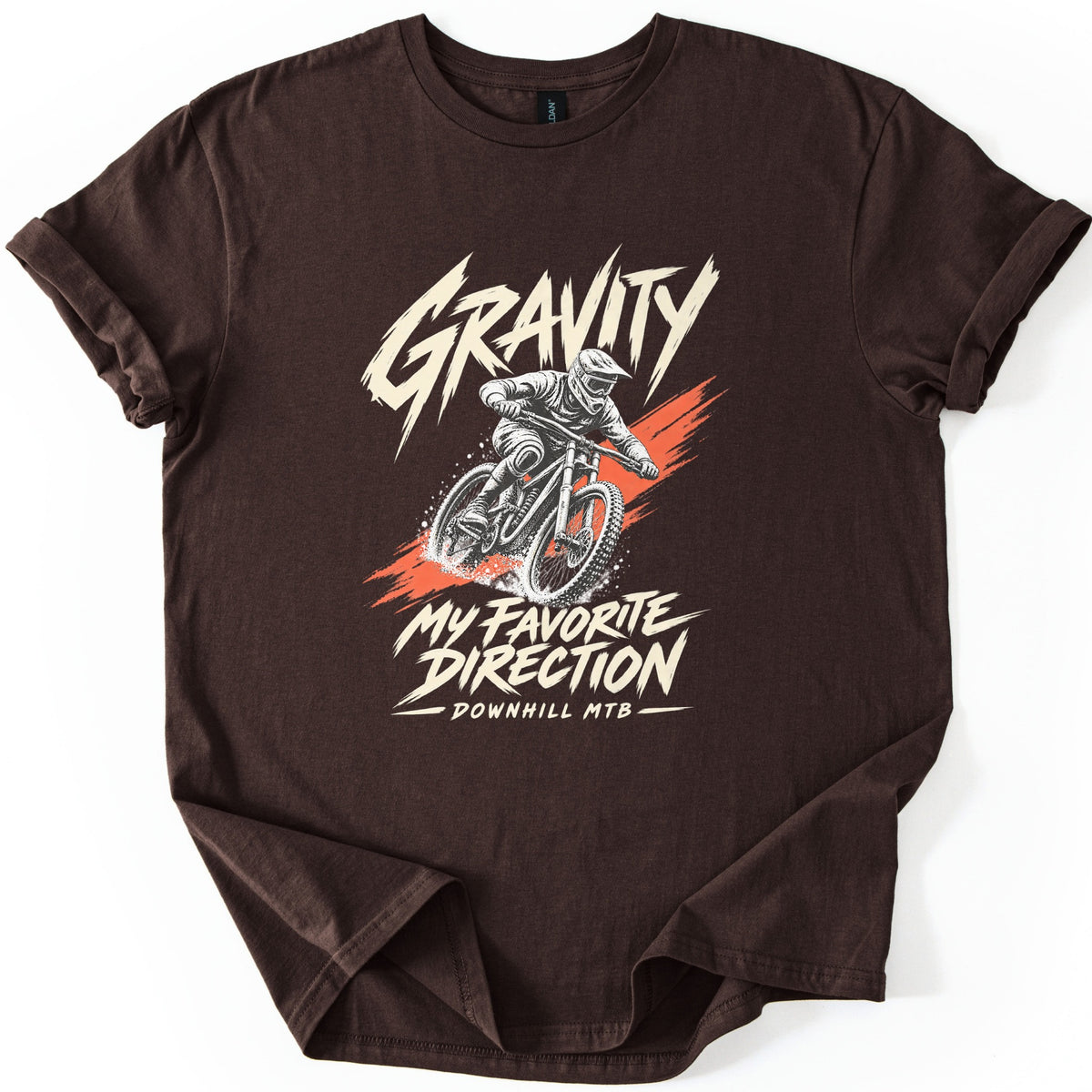 Gravity My Favorite Direction Comfort Colors T-shirt in Dark Chocolate, laid flat. Cream brush lettering reads Gravity above a downhill mountain biker in a full-face helmet spraying dirt over an orange streak, then My Favorite Direction and Downhill MTB. Sleeves cuffed, on white.