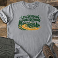Less Skidding More Grinning Comfort Colors T-shirt in Grey, laid flat. Green hand-lettered words Less Skidding More Grinning MTB over a winding gold dirt trail through pine trees toward mountains, with a small smiley face. Olive hiking pants and brown boots lie on weathered wood.