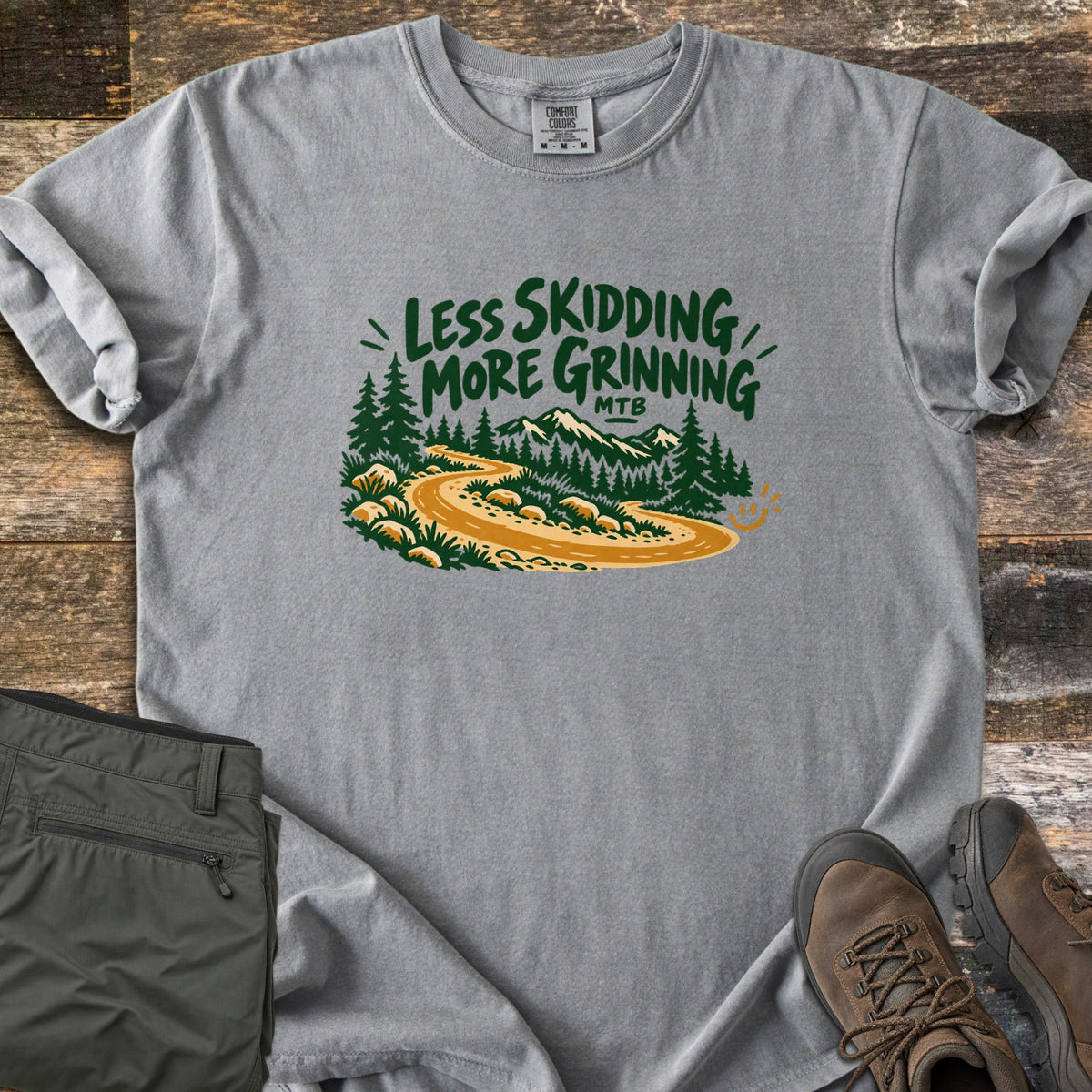 Less Skidding More Grinning Comfort Colors T-shirt in Grey, laid flat. Green hand-lettered words Less Skidding More Grinning MTB over a winding gold dirt trail through pine trees toward mountains, with a small smiley face. Olive hiking pants and brown boots lie on weathered wood.