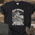Wildlife Photography Because Murder Is Wrong Comfort Colors T-shirt in Black, laid flat. Cream ink shows a wolf on a rocky ledge aiming a long-lens camera at a distant deer by a mountain lake, under Wildlife Photography and above Because Murder Is Wrong. Olive pants and brown hiking boots lie on dark wood.