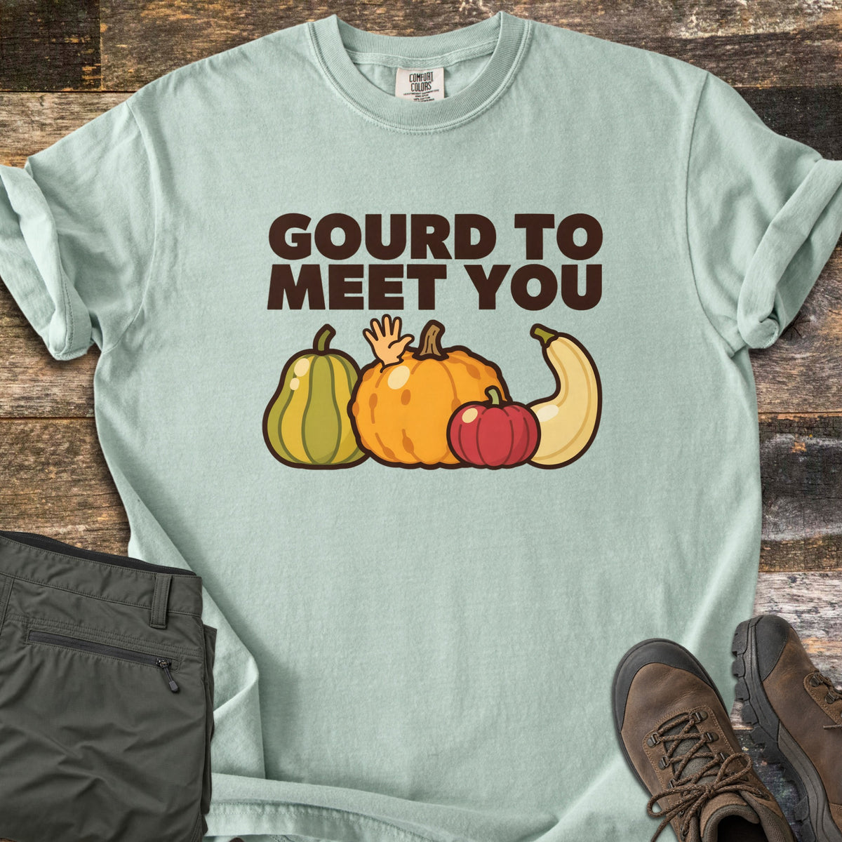 Gourd To Meet You Comfort Colors T-shirt in Bay, laid flat. Dark brown words Gourd To Meet You above cartoon gourds: a green striped squash, an orange pumpkin with a waving hand, a red pumpkin and a cream squash. Olive pants and brown hiking boots sit on wood planks.