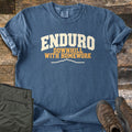 Enduro Downhill With Homework Comfort Colors T-shirt in Denim, laid flat. The word Enduro in large cream arched letters, with Downhill With Homework in orange below and a thin cream mountain ridge line. Sleeves are rolled on a rustic wood surface beside green pants and brown hiking boots.