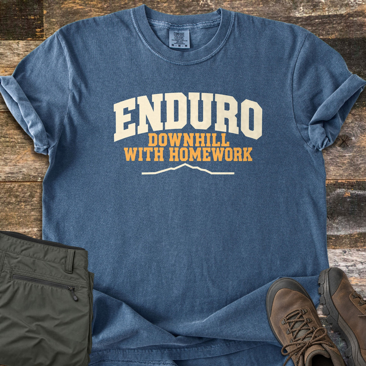 Enduro Downhill With Homework Comfort Colors T-shirt in Denim, laid flat. The word Enduro in large cream arched letters, with Downhill With Homework in orange below and a thin cream mountain ridge line. Sleeves are rolled on a rustic wood surface beside green pants and brown hiking boots.