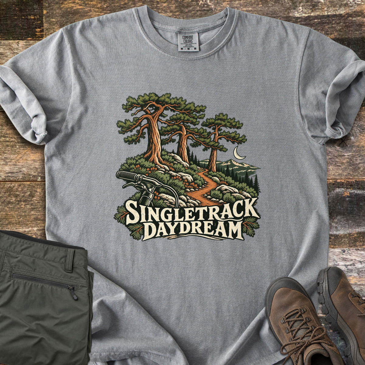Singletrack Daydream Comfort Colors T-shirt in Grey, laid flat. Three tall pines tower over a dirt trail winding past rocks toward mountains and a crescent moon, with bike handlebars in front and the words Singletrack Daydream. Set on wood with olive pants and brown hiking boots.