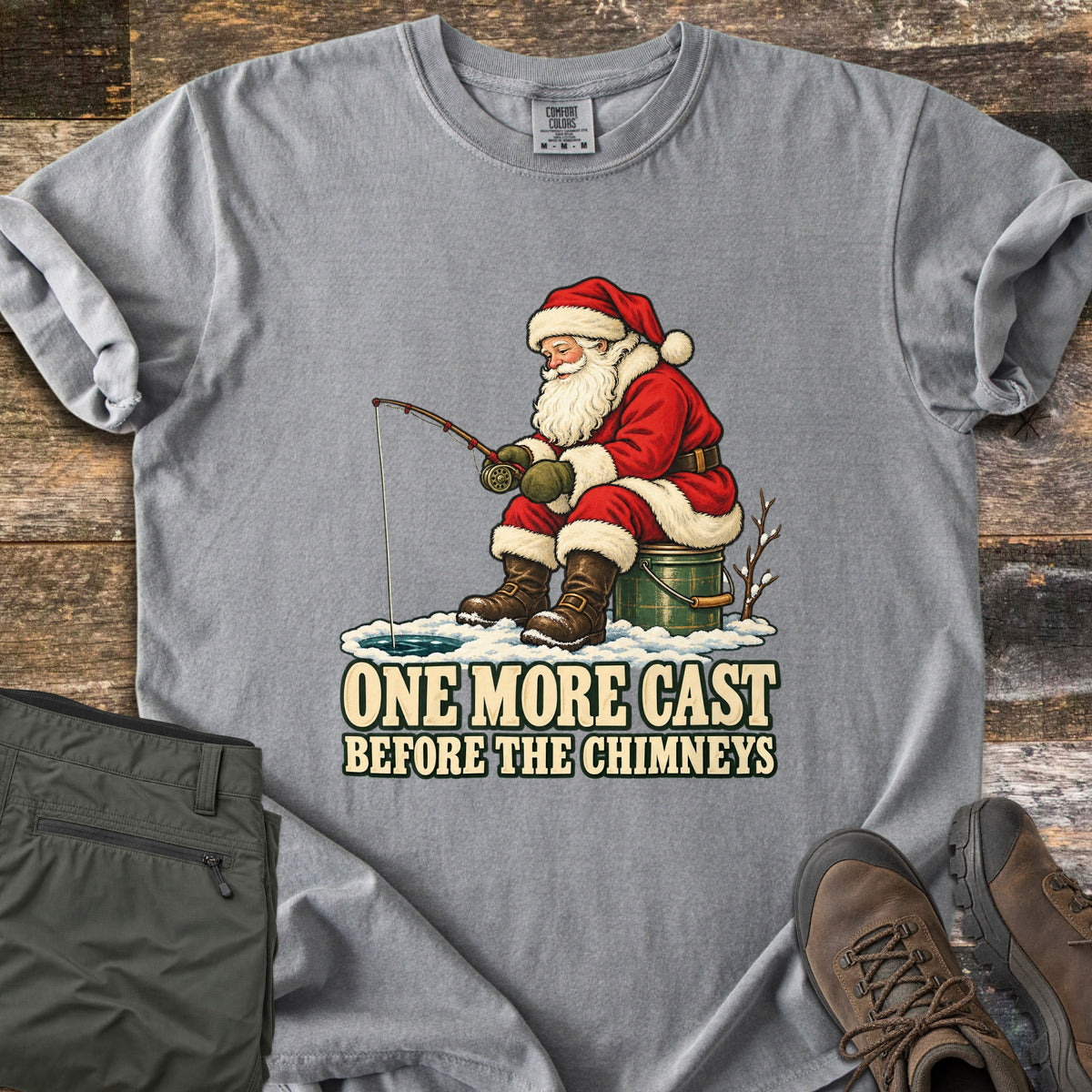 One More Cast Before the Chimneys Comfort Colors T-shirt in Grey, laid flat. Santa in his red suit sits on a green bucket, ice fishing over a hole in the snow, above the words One More Cast Before the Chimneys. Olive pants and brown hiking boots lie beside it on weathered wood.