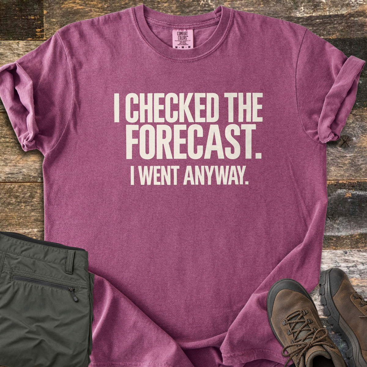 Checked The Forecast Comfort Colors T-shirt in Berry, laid flat. Off-white block letters read I CHECKED THE FORECAST. I WENT ANYWAY. The sleeves are cuffed, with folded olive hiking pants and brown hiking boots beside it on weathered wood planks.