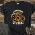 I Think He Wants Uppies Comfort Colors T-shirt in Black, laid flat. A cartoon brown bear with wild eyes, a lolling tongue, and raised clawed paws sits between the words I Think He Wants Uppies! Shown on wood planks beside folded green hiking pants and brown hiking boots.