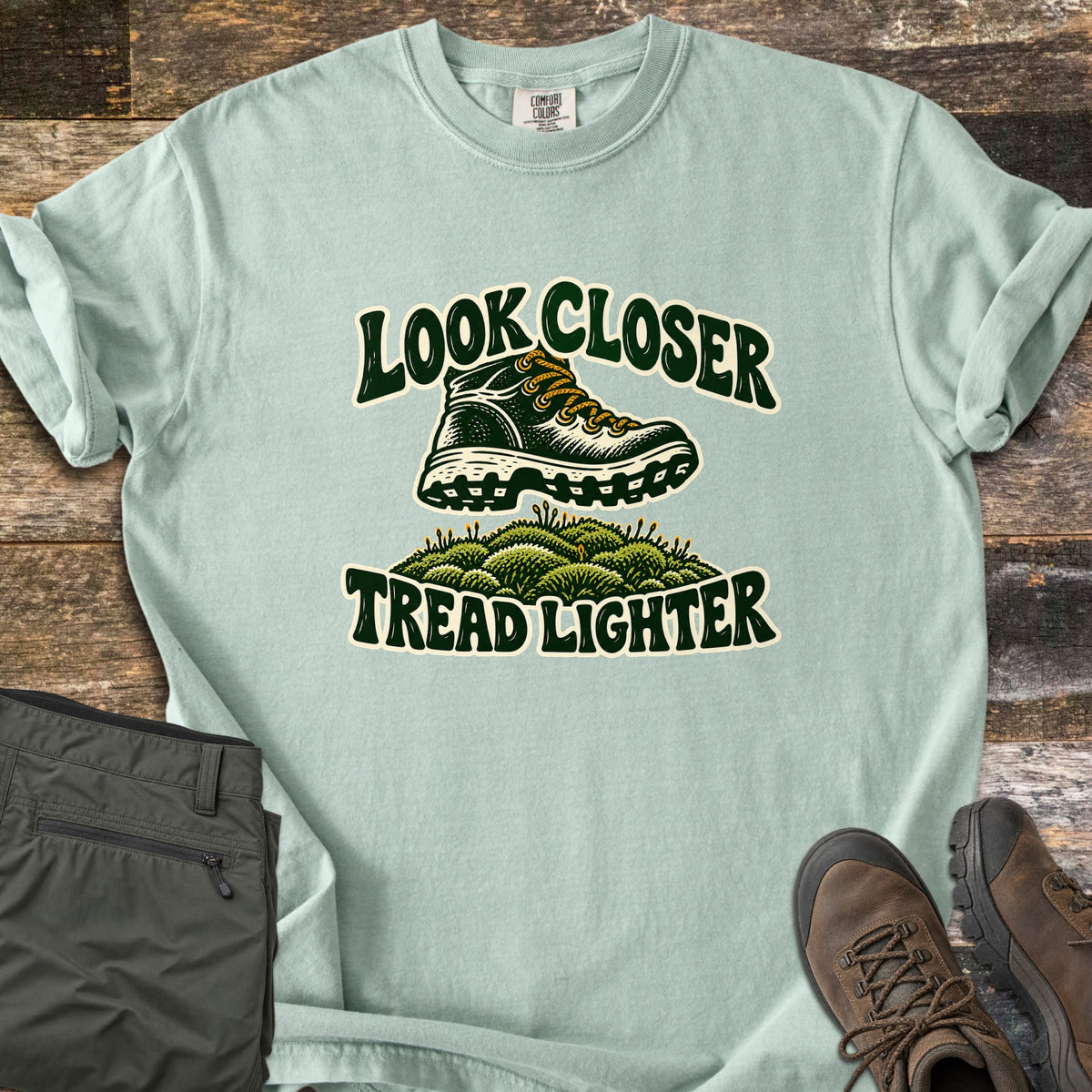 Look Closer Tread Lighter Comfort Colors T-shirt in Bay, laid flat. A green hiking boot with yellow laces steps above a mound of moss between the words Look Closer and Tread Lighter. Sleeves rolled, on weathered wood with folded olive hiking pants and brown boots.