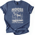 Moose Traffic Control Comfort Colors T-shirt in Heather Navy, laid flat. A cream sketch of a bull moose walking across a road between pine trees, under the words Moose Traffic Control and above Yield to Antlers. The sleeves are cuffed, on a plain white background.