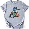 Kingfisher Waterfall Comfort Colors T-shirt in Sport Grey, laid flat. A painted kingfisher with a blue crest perches on a branch, its body filled with a waterfall pouring into a turquoise pool among rocks and pine trees. No words. Sleeves rolled, on a white background.