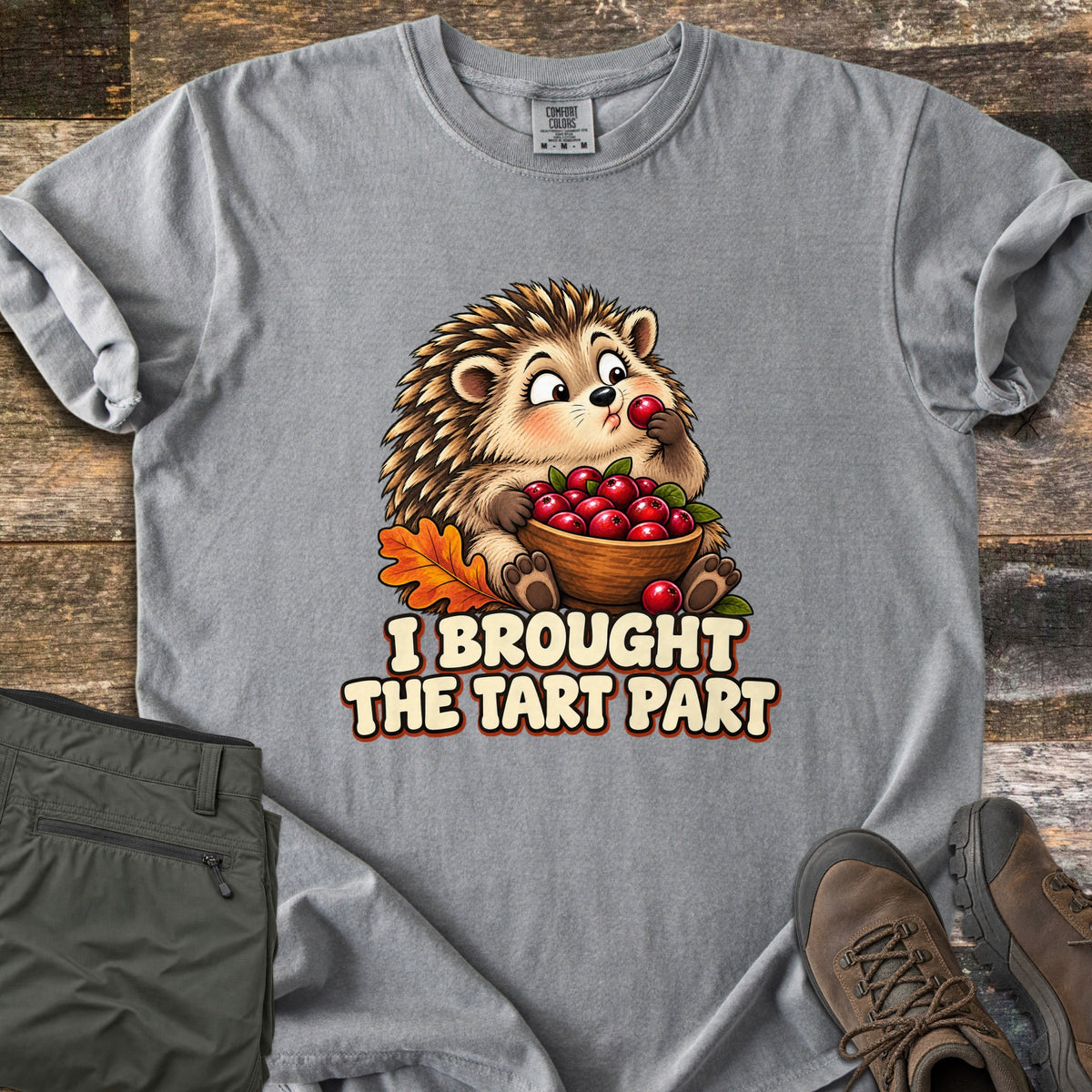 I Brought the Tart Part Comfort Colors T-shirt in Grey, laid flat. A cartoon hedgehog nibbles a cranberry while holding a wooden bowl of cranberries beside an orange oak leaf, above the words I Brought the Tart Part. Olive pants and brown hiking boots sit on a wood background.