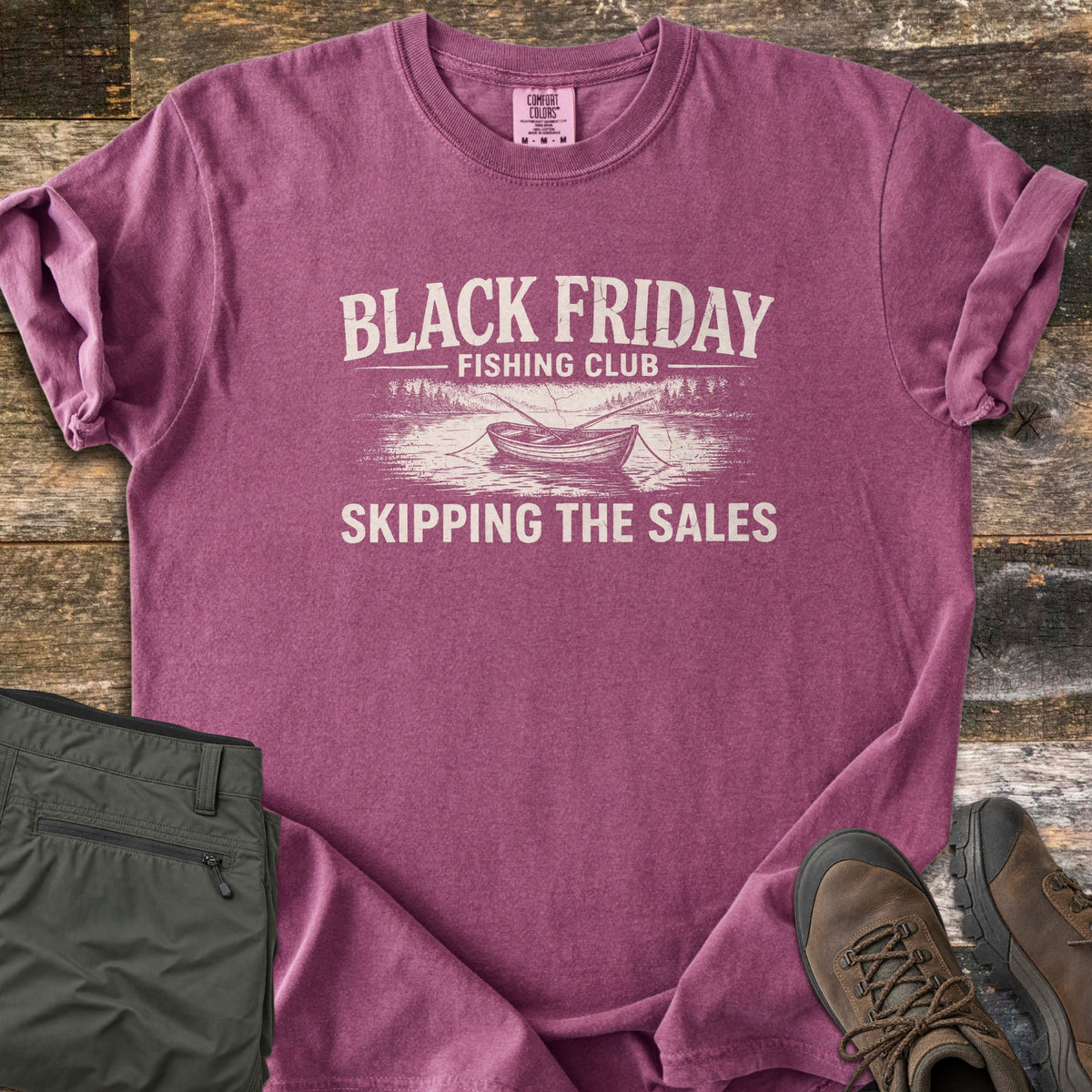 Black Friday Fishing Club Comfort Colors T-shirt in Berry, laid flat. A distressed cream print shows the words Black Friday Fishing Club above a rowboat with oars on a lake lined with pine trees, and Skipping the Sales below. Olive pants and brown hiking boots sit beside it on wood planks.