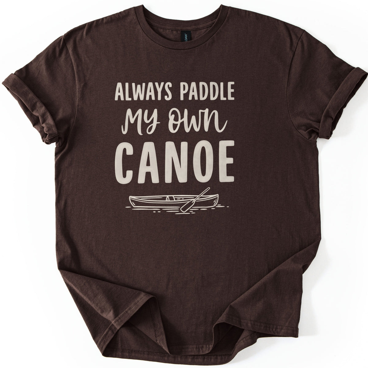 Paddle My Own Canoe Comfort Colors T-shirt in Dark Chocolate, laid flat. Cream text reads Always Paddle My Own Canoe above a line drawing of a canoe with a paddle on rippling water. The shirt has rolled sleeves and lies on a white surface.