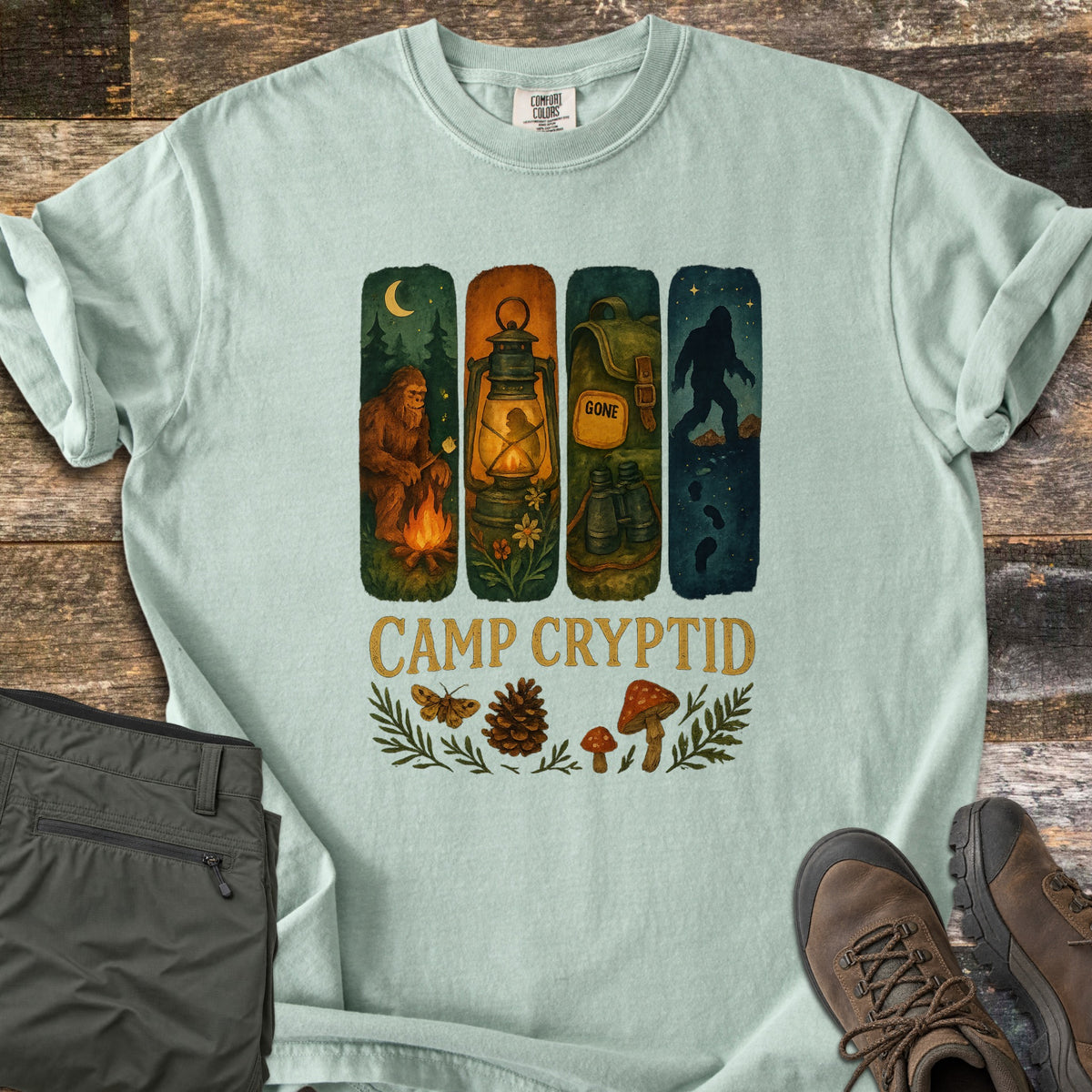 Camp Cryptid Big Foot Comfort Colors T-shirt in Bay, laid flat. Four painted panels show Bigfoot at a campfire, a lantern, a backpack marked GONE with binoculars, and Bigfoot under stars, above CAMP CRYPTID, a moth, pinecone and mushrooms. Olive pants and hiking boots lie on wood.