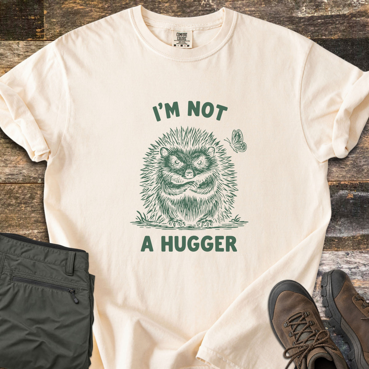 I'm Not A Hugger Comfort Colors T-shirt in Ivory, laid flat. A green sketch of a grumpy hedgehog with crossed arms and a butterfly beside it, between the words I'M NOT A HUGGER. Shown on wood boards with folded olive hiking pants and brown hiking boots.