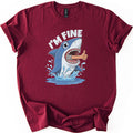 Shark I'm Fine Comfort Colors T-shirt in Maroon, laid flat. A cartoon blue shark leaps from splashing water with a human arm giving a thumbs up sticking out of its open mouth, under the words I'M FINE in white. The shirt has rolled sleeves on a white background.