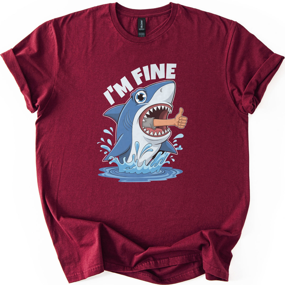 Shark I'm Fine Comfort Colors T-shirt in Maroon, laid flat. A cartoon blue shark leaps from splashing water with a human arm giving a thumbs up sticking out of its open mouth, under the words I'M FINE in white. The shirt has rolled sleeves on a white background.