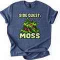 Side Quest Moss Comfort Colors T-shirt in Heather Navy, laid flat. A pixel-art tree stump covered in green moss and ferns, with two tiny spore stalks and sparkles, sits between the words Side Quest: and Moss in cream pixel letters. White background, rolled sleeves.