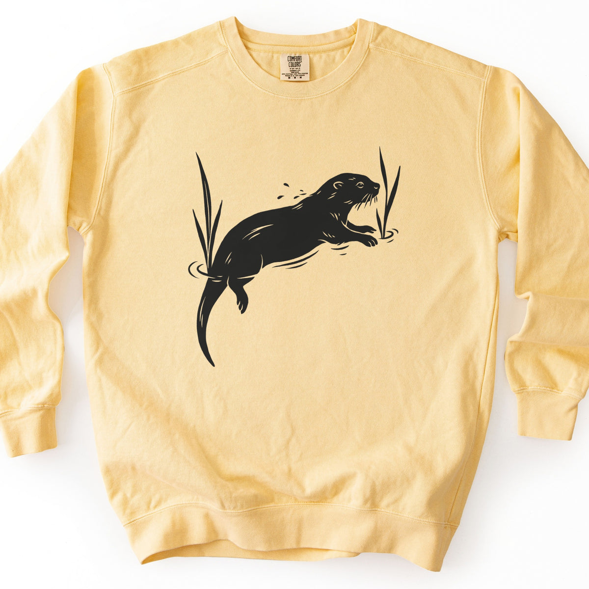 Swimming Otter Comfort Colors crewneck sweatshirt in Butter, laid flat. A black block-print style otter swims with its head raised, splashing droplets, between two tall reeds with ripples around them. No words are printed. The sweatshirt lies on a white background.