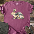 Snowshoe Hare Birch Woods Comfort Colors T-shirt in Berry, laid flat. A cream snowshoe hare sits in profile, its body filled with a snowy birch forest, a winding path and distant mountains; no words. Sleeves rolled, on weathered wood with olive pants and brown hiking boots.