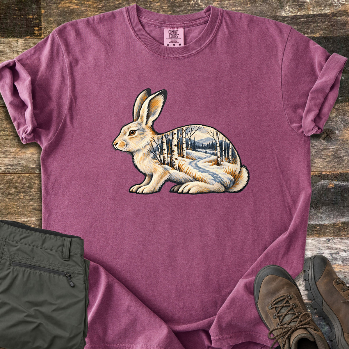 Snowshoe Hare Birch Woods Comfort Colors T-shirt in Berry, laid flat. A cream snowshoe hare sits in profile, its body filled with a snowy birch forest, a winding path and distant mountains; no words. Sleeves rolled, on weathered wood with olive pants and brown hiking boots.