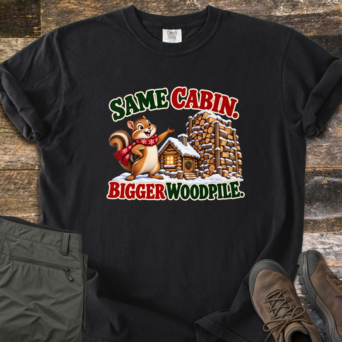 Same Cabin. Bigger Woodpile. Comfort Colors T-shirt in Black, laid flat. A chipmunk in a red snowflake scarf gestures at a snowy log cabin beside a towering woodpile, between the words Same Cabin. Bigger Woodpile. Shown on weathered wood with olive pants and brown hiking boots.
