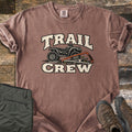 Trail Crew Comfort Colors T-shirt in Espresso, laid flat. The words TRAIL CREW in cream letters frame a black ATV over leafy ground with an orange bow saw cutting a log. Olive hiking pants and brown hiking boots sit beside it on weathered wood boards.