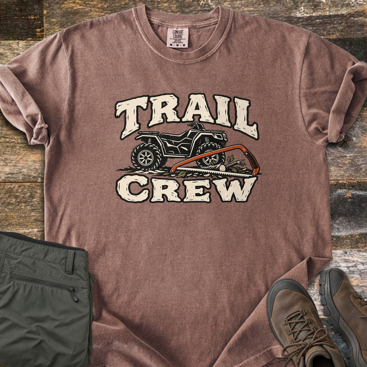 Trail Crew Comfort Colors T-shirt in Espresso, laid flat. The words TRAIL CREW in cream letters frame a black ATV over leafy ground with an orange bow saw cutting a log. Olive hiking pants and brown hiking boots sit beside it on weathered wood boards.
