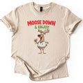 Moose Down And Enjoy Comfort Colors T-shirt in Sand, laid flat. Red words Moose Down above green & Enjoy, over a grinning cartoon moose in a white short-sleeve shirt holding a glass of orange drink, hand on hip. Sleeves are cuffed on a white background.