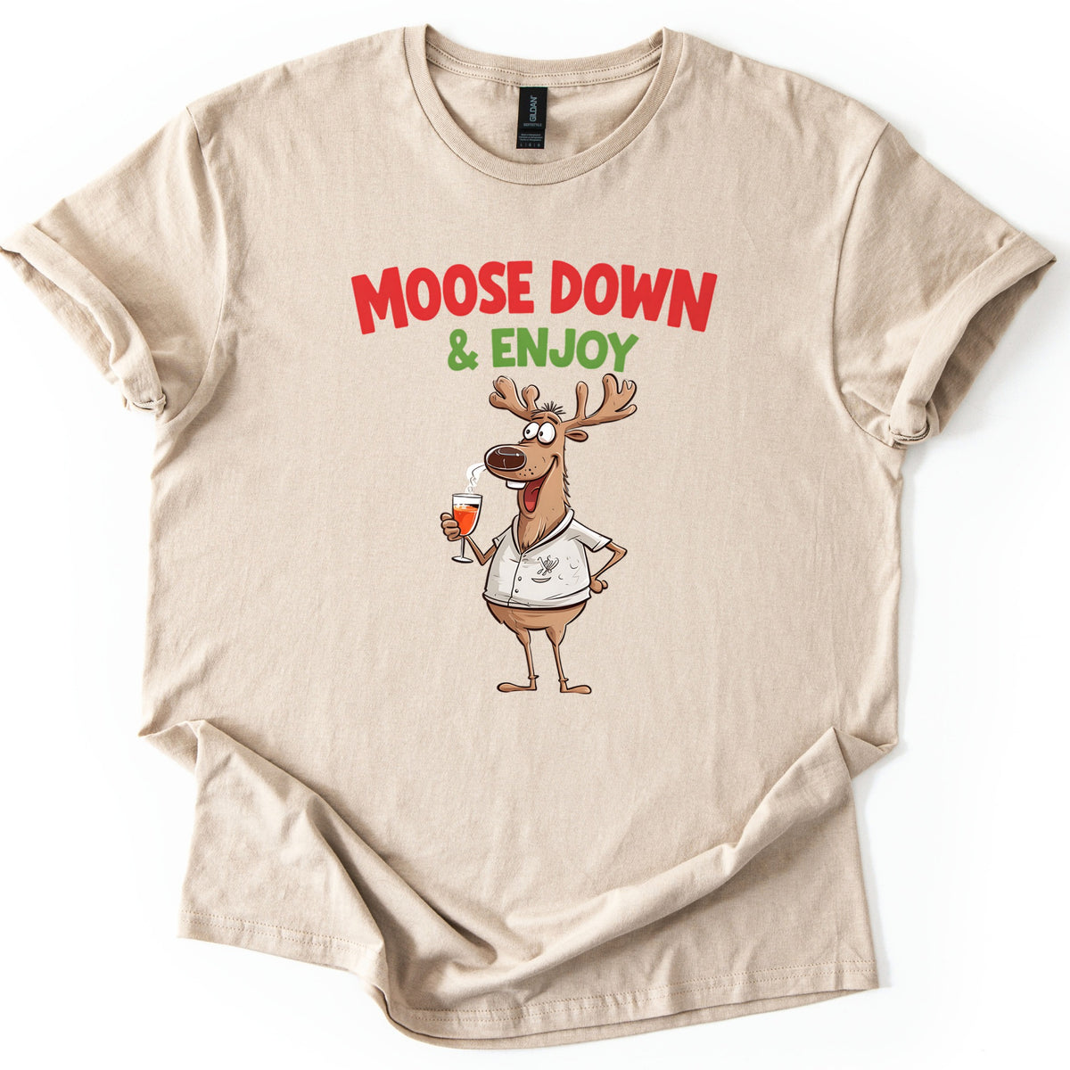 Moose Down And Enjoy Comfort Colors T-shirt in Sand, laid flat. Red words Moose Down above green & Enjoy, over a grinning cartoon moose in a white short-sleeve shirt holding a glass of orange drink, hand on hip. Sleeves are cuffed on a white background.